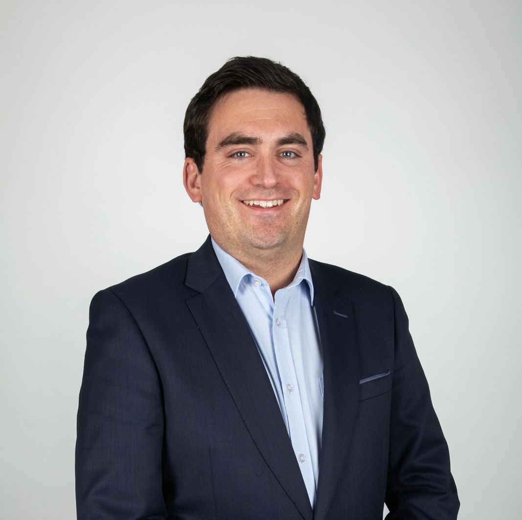 picture of Labour Party candidate Shane Folan smiling, wearing a navy blazer and light blue shirt with top button open